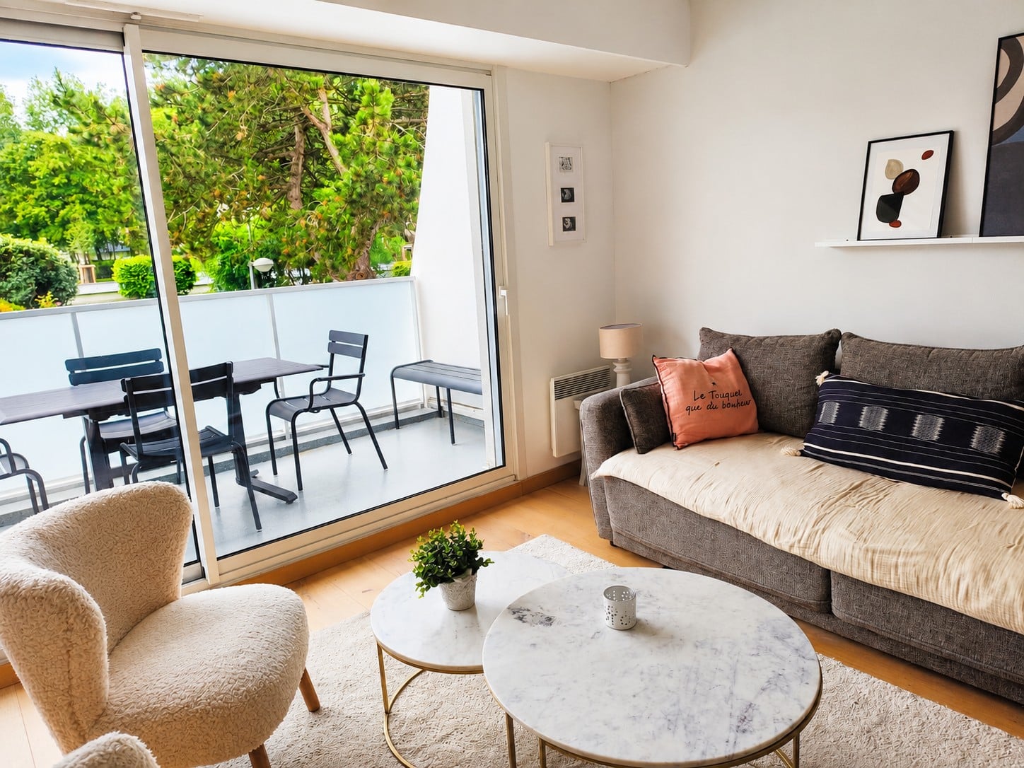 Séjour lumineux ouvert sur un balcon : fauteuil bouclette, tables basses en marbre, canapé garni de coussins et pins derrière la baie vitrée