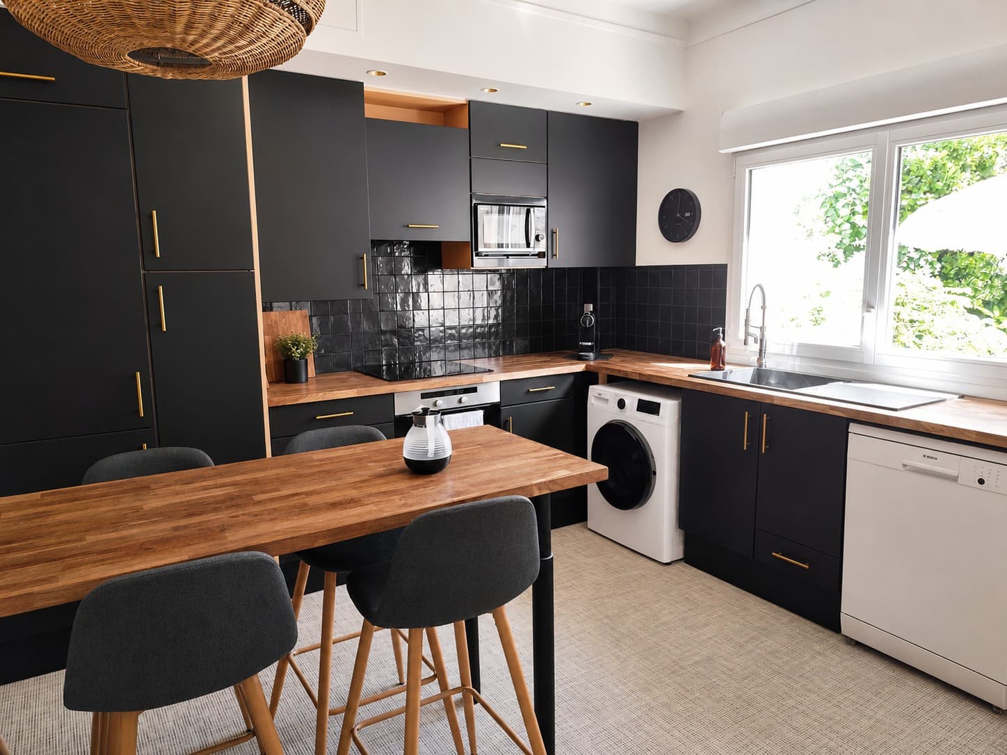 Cuisine aux façades noires et poignées dorées, plan de travail et table haute en bois, lumière naturelle et vue sur la verdure