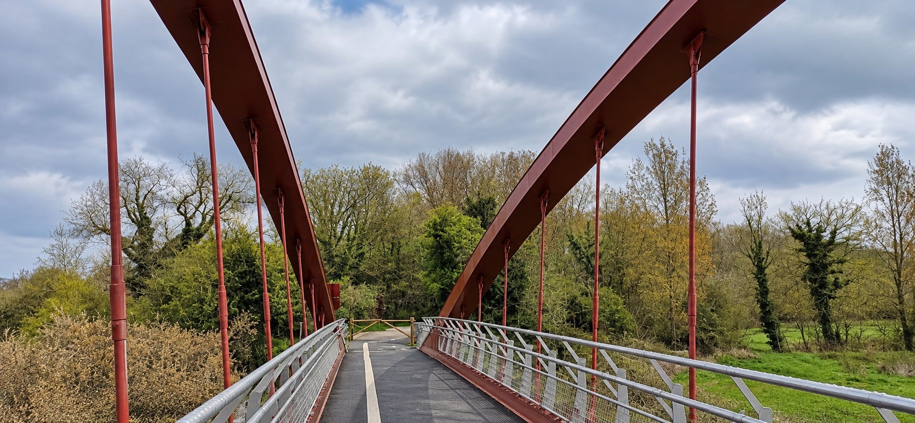 Passerelle piétonne et cyclable aux arches rouges, menant vers des arbres et des prairies