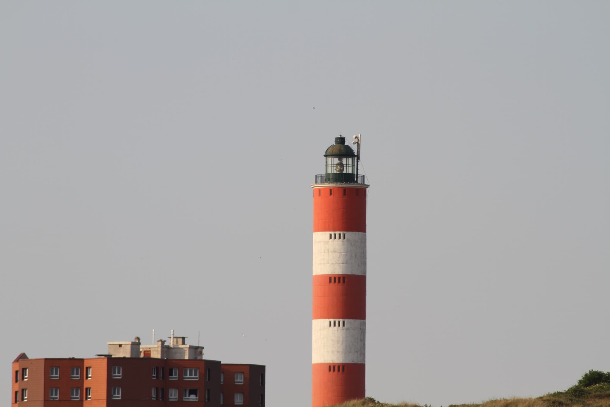 Phare à bandes rouges et blanches au-dessus des dunes, à côté d’un immeuble de brique