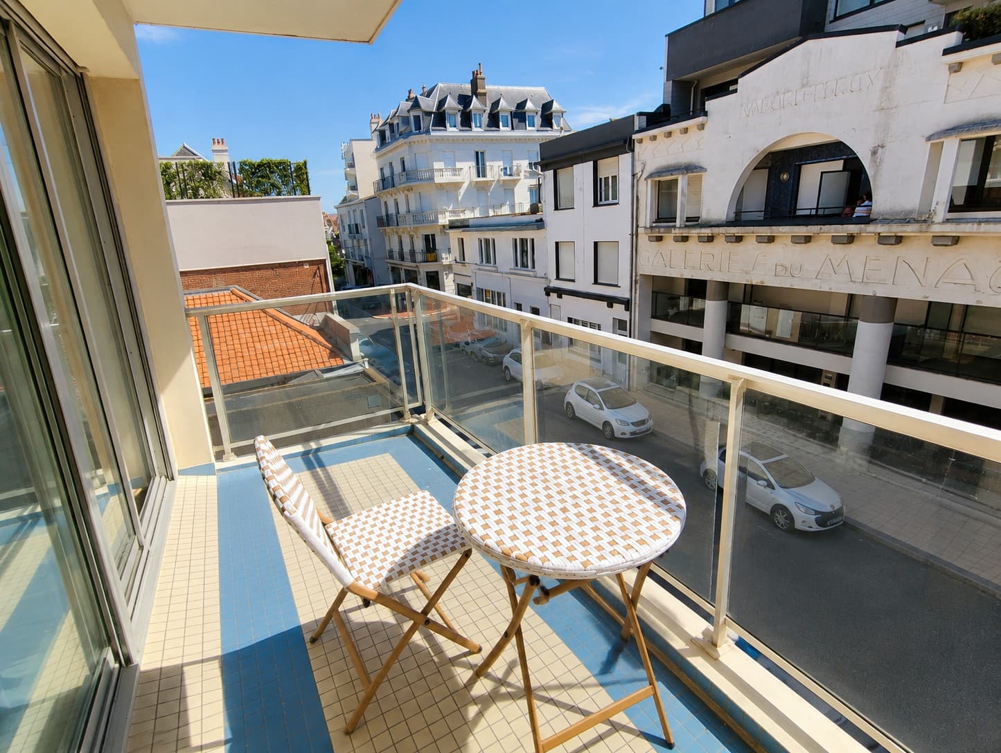 Balcon d’appartement meublé d’une table et d’une chaise de bistrot, au-dessus d’une rue bordée d’immeubles blancs aux toits d’ardoise