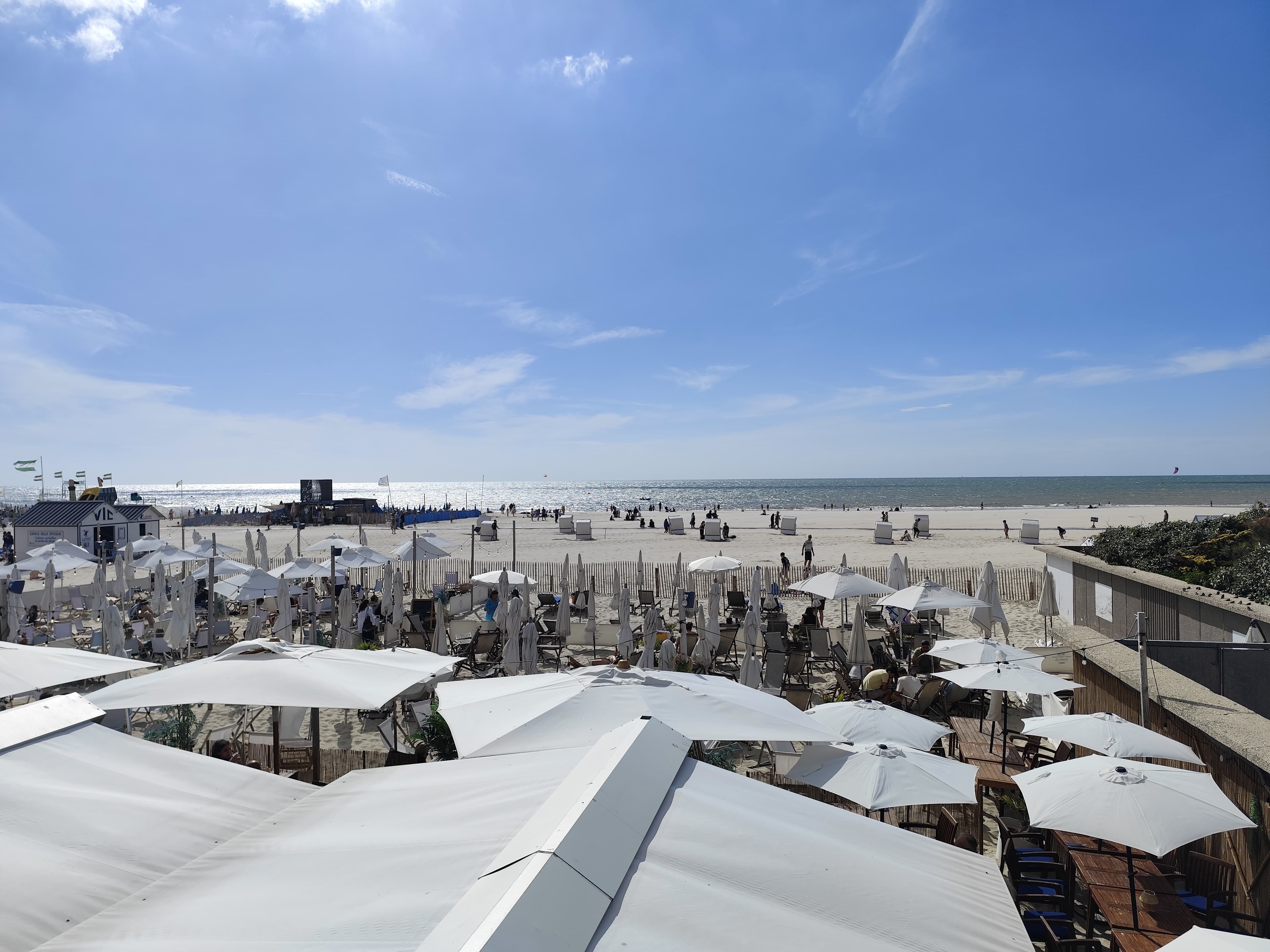 Terrasses de plage sous des parasols blancs, devant la grande plage de sable et la mer à l’horizon
