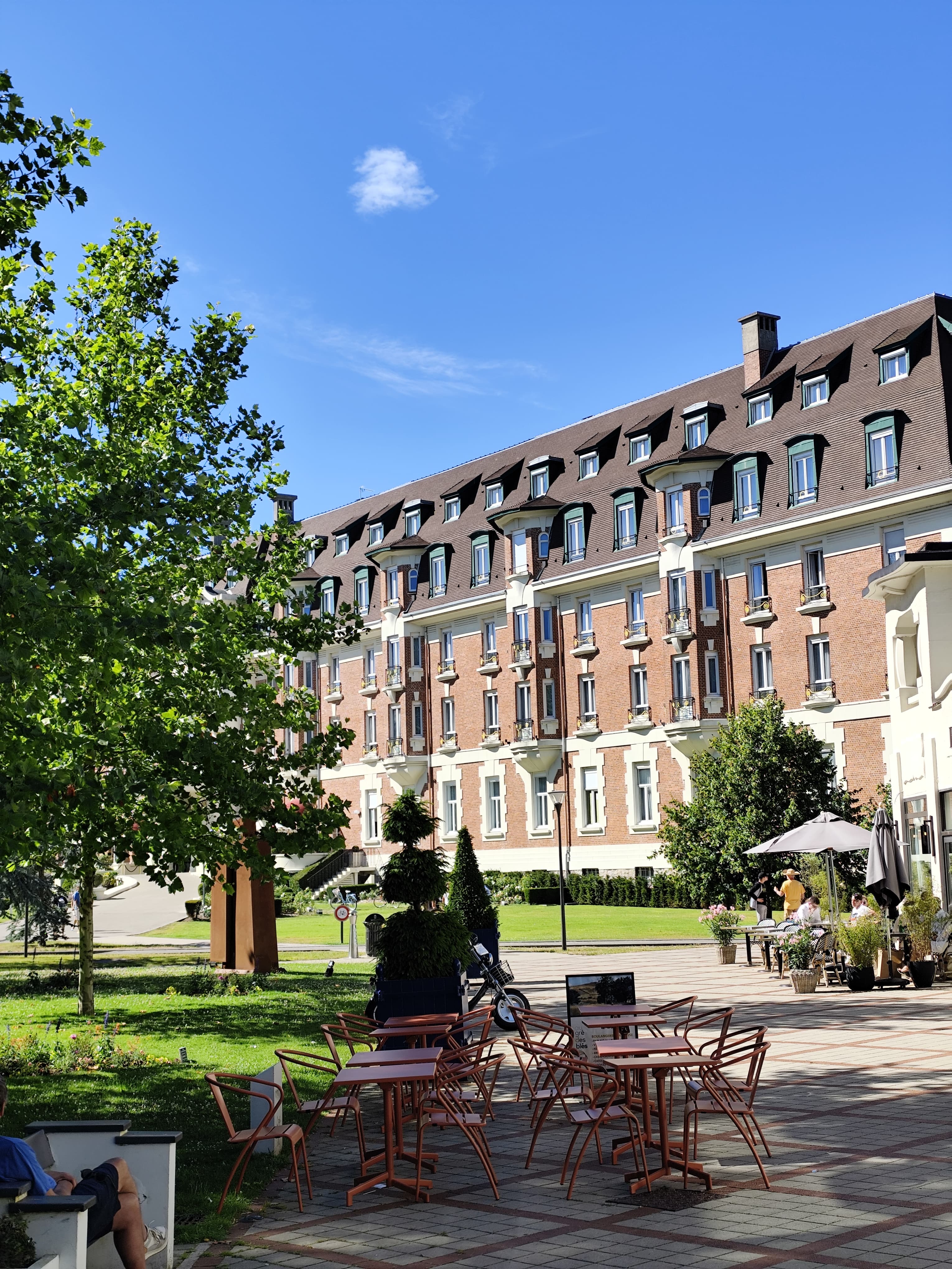 Grand bâtiment de brique aux toits mansardés et aux balcons blancs, derrière un jardin arboré et une terrasse de café