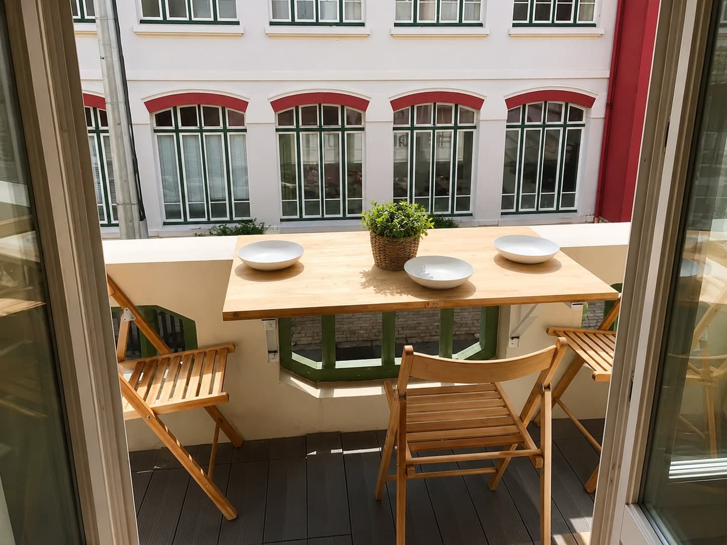 Balcon en bois clair, table dressée, face à une façade blanche aux fenêtres cintrées soulignées de brique et menuiseries vertes