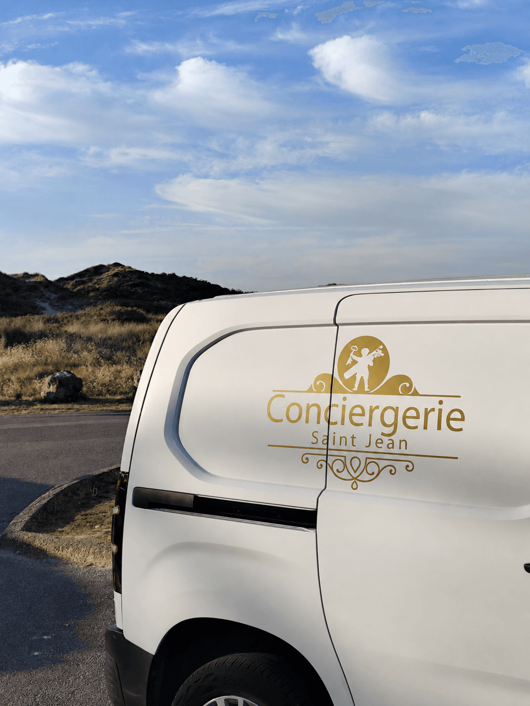 Véhicule utilitaire blanc marqué du logo doré de la Conciergerie Saint-Jean, garé devant les dunes